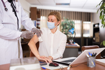 Doctor in white coat and protective mask giving vaccine injection to woman manager