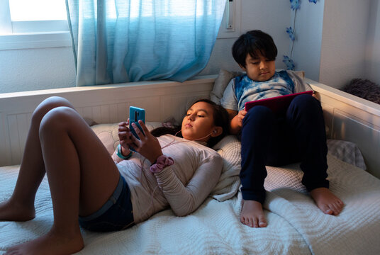 Hispanic Brother and sister at home with devices on the bed.