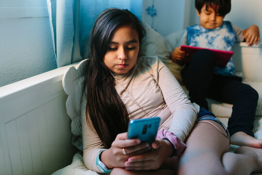 Hispanic Brother and sister at home with devices on the bed.