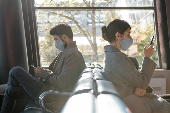 Travelers in masks using smartphones against window in airport