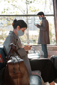 Woman in mask using smartphone near man in waiting room of airport