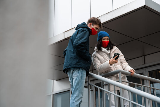 Multiethnic man and woman in masks browsing smartphone outside building