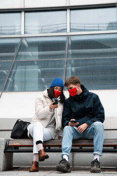 Young couple in masks using smartphones on bench