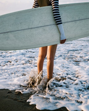 Photo of the lags of a surfer girl who stands with a surfboard on the shoreline. Surfing on Bali island 