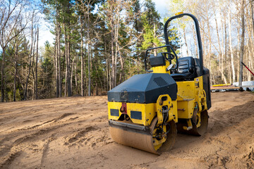 Closeup of yellow compactor roller, a machined used for compaction of soil, asphalt surface, gravel and sand. Viewed on an empty, tree lined building lot, ready for new home construction.
