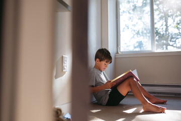 Little boy reading on the floor at home near window.
