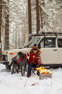 Female traveler with dog warming near fire in winter forest