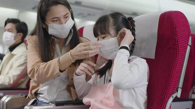 Asian mother helping her daughter wear face mask during seating on plane travel abroad during covid pandemic for prevent coronavirus infection. Transportation business or health care concept.