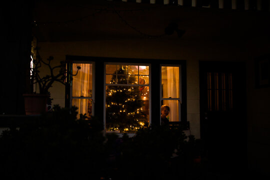Outside view of young child peering through window while father decorates tree inside
