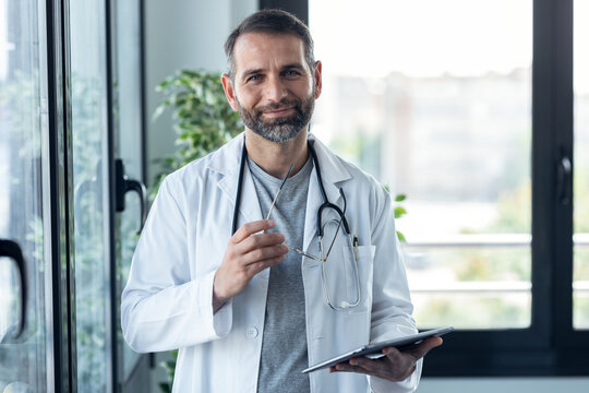 Handsome mature male doctor workinh while holding digital tablet looking at camera in the medical consultation.