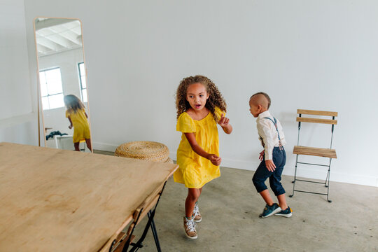 Young biracial brother and sister run around room with wood table and mirror