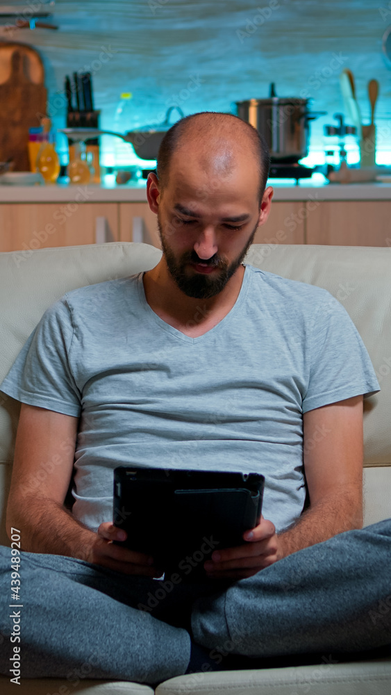Poster tired man sitting alone on couch while browsing on internet using tablet computer with modern techno