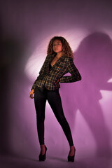 vertical portrait of a curly model wearing a jacket, black pants and high heel shoes in a purple backgroung
