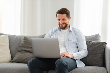 Happy caucasian man uses laptop computer for video call while sitting on sofa at home, smiling caucasian man chatting with friends or family via video conference on social networks