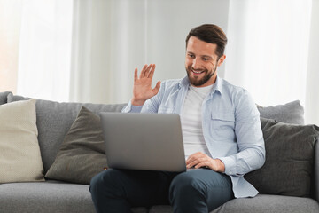 Happy caucasian man uses laptop computer for video call while sitting on sofa at home, smiling caucasian man chatting with friends or family via video conference on social networks, greeting gesture