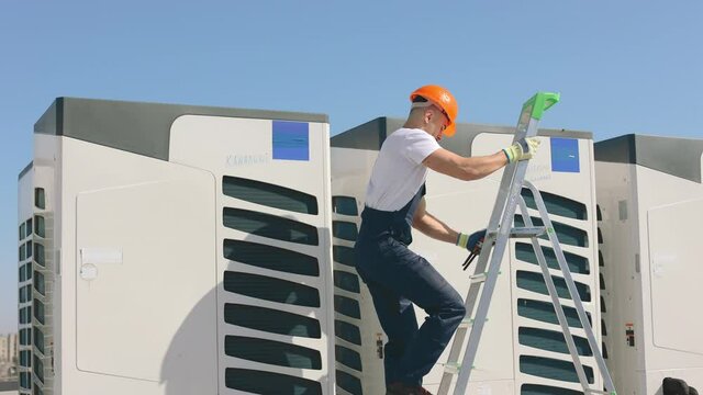 The young engineer is climbing up the ladder and twisting the screws with a pipe wrench. He is maintaining the air conditioning system. He is wearing work clothes and a hard hat. Nice sunny day. 4K