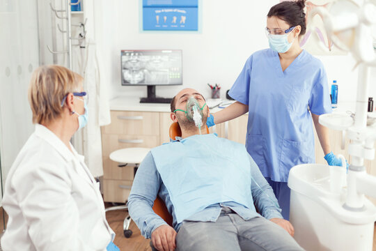 Physician assistant putting oxigen mask before tooth surgery standing on dental chair in stomatology cabinet. Senior dentist doctor and nurse working in modern office wearing mask and gloves