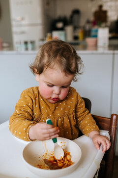 Baby girl with polkadot dress eating pasta in a high chair