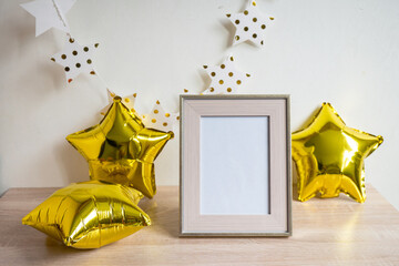 Portrait white picture frame mockup on wooden table. Gold baloons, birstday concept. White wall...