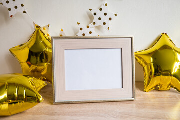 Portrait white picture frame mockup on wooden table. Gold baloons, birstday concept. White wall...