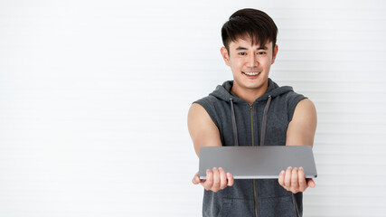 Asian handsome fit smiling man stand hand over a laptop computer wearing sleeveless t-shirt gray...