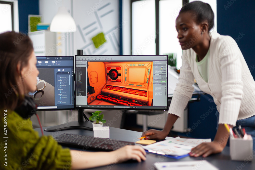 Poster multiethnic women game designer looking at computer with dual displays working together at project i