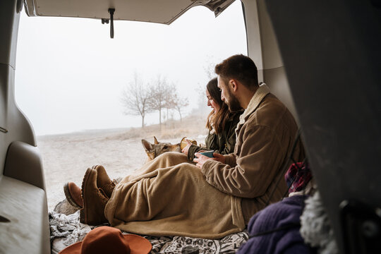 Young couple in a camper van in nature during winter with cute dog having warm coffe
