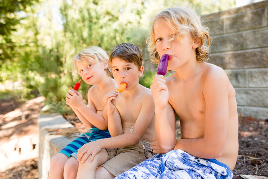Shirtless boys sit on a stone wall eating popsicles