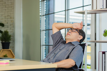 Asian businessman tired overworked he relaxing with closed eyes and hands behind his head on the desk. senior man with eyeglasses lying asleep on table at his working place
