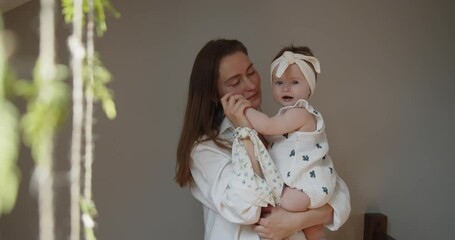 Mother and daughter spend time at home together. Young mom holds her little baby girl close to her standing under the window playing with hanging decoration. Parent care and child safety.