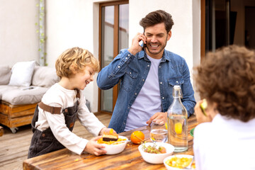 Cheerful father talking on phone during breakfast on terrace
