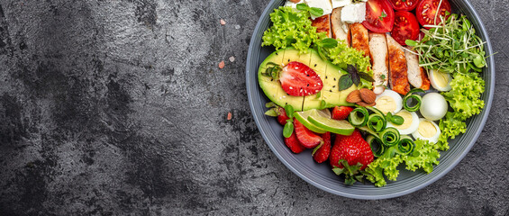Healthy dinner. Buddha bowl lunch with grilled chicken and avocado, feta cheese, quail eggs, strawberries, nuts and lettuce on gray background. Long banner format. top view