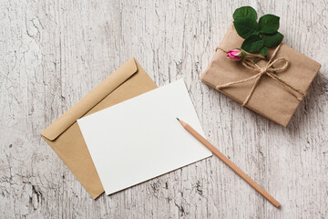 Mockup blank white greeting card or invitation, gift box wrapped with kraft paper and pink rose bud on gray table. Blank sheet of paper for text. Holiday concept. Top view.