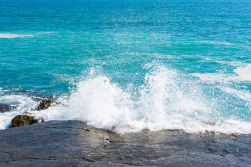 White wave of the turquoise sea breaks on a stone on the shore on a bright sunny day