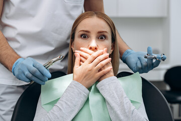 Attractive, young woman covers her mouth with her hands in a dental chair. The woman is afraid to...