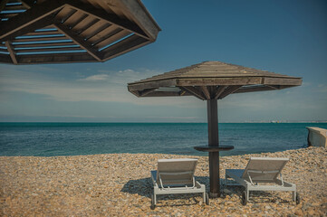 two white sun loungers under a wooden beach umbrella on a rocky beach in summer on a clear day without people