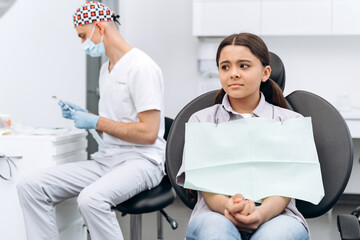 Sad little girl in a dental chair. Against the background of blurred doctor prepares working tools...