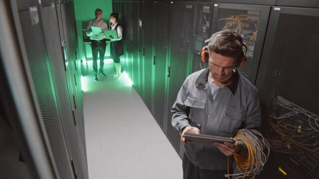 High angle of Caucasian male technician wearing safety ear muffs and worker clothing, using tablet computer in server room