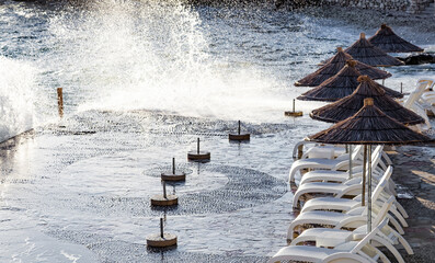 Beach umbrellas and sunbeds against the sea waves in a strong storm. Swimming is dangerous and strongly prohibited, spoiled vacation, bad weather. Nobody on the beach.