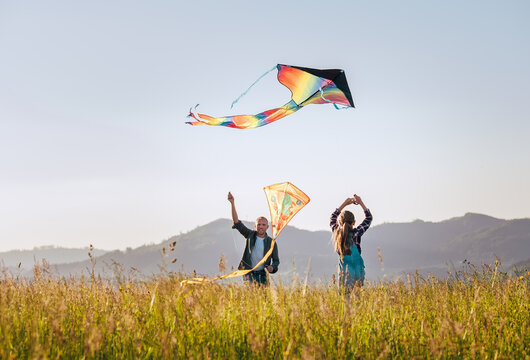 Daughter with smiling father while they flying a colorful kites on the high grass meadow in the mountain fields. Warm family moments or outdoor time spending concept image.