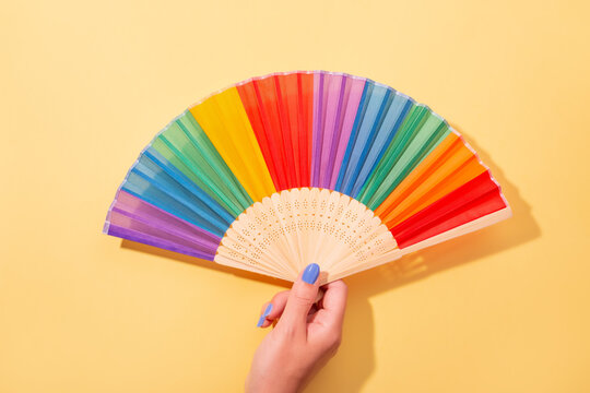 Woman with bright blue manicure holding fan in rainbow colors on yellow background, top view