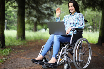 Young disabled woman in wheelchair waving hand at laptop screen in park