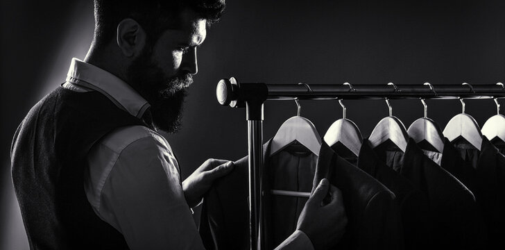 Male suits hanging in a row. Men clothing, boutiques. Tailor, tailoring. Stylish men's suit. Man suit, tailor in his workshop. Handsome bearded fashion man in classical costume suit. Black and white
