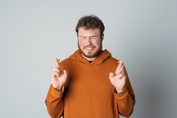 Hopeful bearded handsome young man cross fingers, close eyes and plead about something, standing over gray background