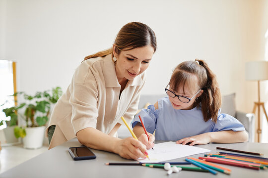 Portrait of cute girl with down syndrome studying at home with mother helping her in cozy interior, copy space