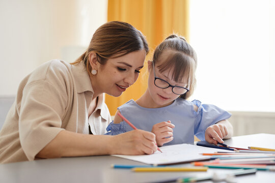 Portrait of cute girl with down syndrome studying at home with caring mother helping her in cozy interior