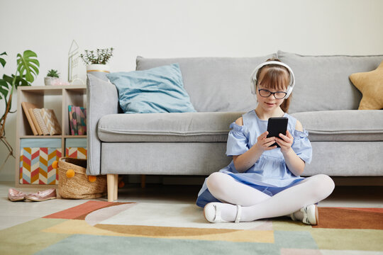 Candid full length portrait of teenage girl with down syndrome listening to music at home while sitting on floor in cozy interior, copy space