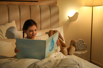Little girl reading book in bedroom lit by night lamp