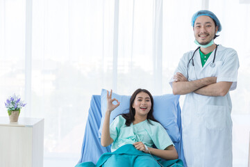 Positve good looking male doctor with white coat is smiling, standing near happy young female patient showing OK sign with hospital clothes on hospital bed in hospital.