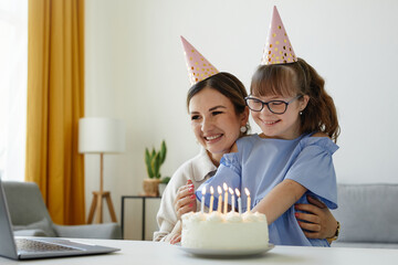 Portrait of happy mother and daughter celebrating birthday online and blowing candles by laptop